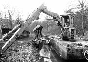 Working immediately east of the Iron Aqueduct over the railway. We are using the hydraulic take-off on the machine to power a jack hammer with piling head to drive piles along the side of the canal bed. This is the first of a great many 4ft steel piles we inserted.